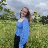 Casey stands in a field of wildflowers with storm clouds in the back.She has light brown hair and a blue shirt.