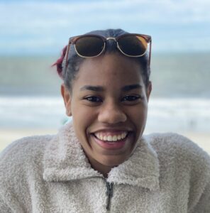 Daisja grins with a beach background, wearing a fluffy jacket and cool sunglasses.