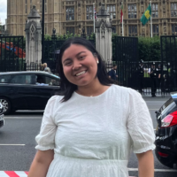 Stephanie, with a black bob and a white dress, smiles in front of Buckingham Palace.