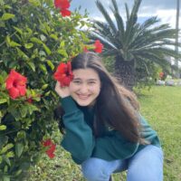 Sarah, a white woman with dark hair, kneels and grins by a hibiscus bush with one of the bright red flowers "tucked" behind her ear.